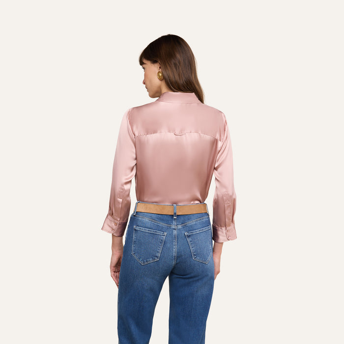 A woman with long brown hair is seen from behind, wearing the Dani Silk Blouse in shiny pink 100 silk charmeuse with rolled-up sleeves and blue jeans, standing against a plain white background.