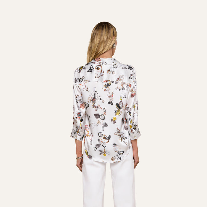 A woman stands facing away, wearing the Dani Silk Blouse—a white silk button-down adorned with a colorful butterfly print—and white pants, set against a plain light background.
