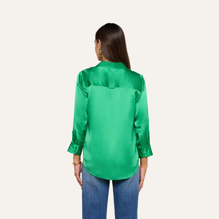 A woman with long brown hair is shown from the back, wearing a bright green Dani Silk Blouse with long sleeves and blue jeans, standing against a plain light background.