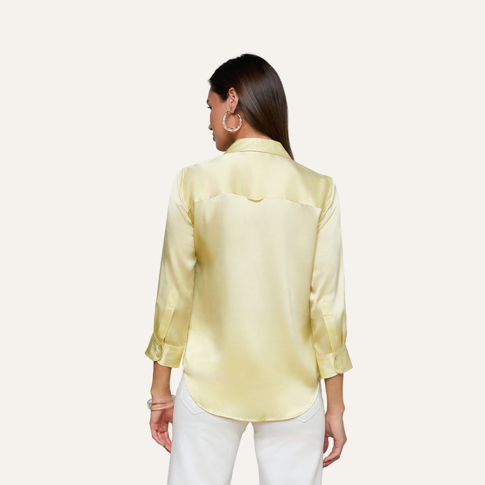 A woman with long brown hair is shown from the back against a plain light background, wearing the Dani Silk Blouse—a light yellow satin top with three-quarter sleeves—and white pants.