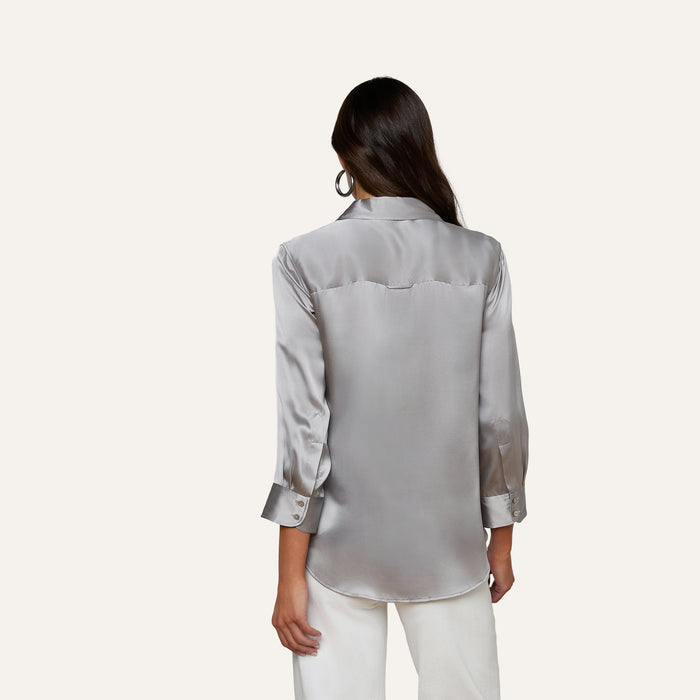 A woman with long dark hair, wearing the Dani Silk Blouse—a shiny silver silk button-down with long sleeves—paired with white pants, stands facing away from the camera against a plain light background.