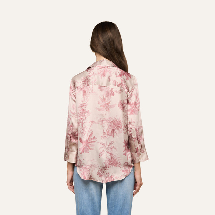 A woman with long brown hair, viewed from behind, is wearing the Dani Silk Blouse—a pink satin button-down featuring a subtle floral print and long sleeves. She pairs it with light blue jeans against a plain, light-colored background.