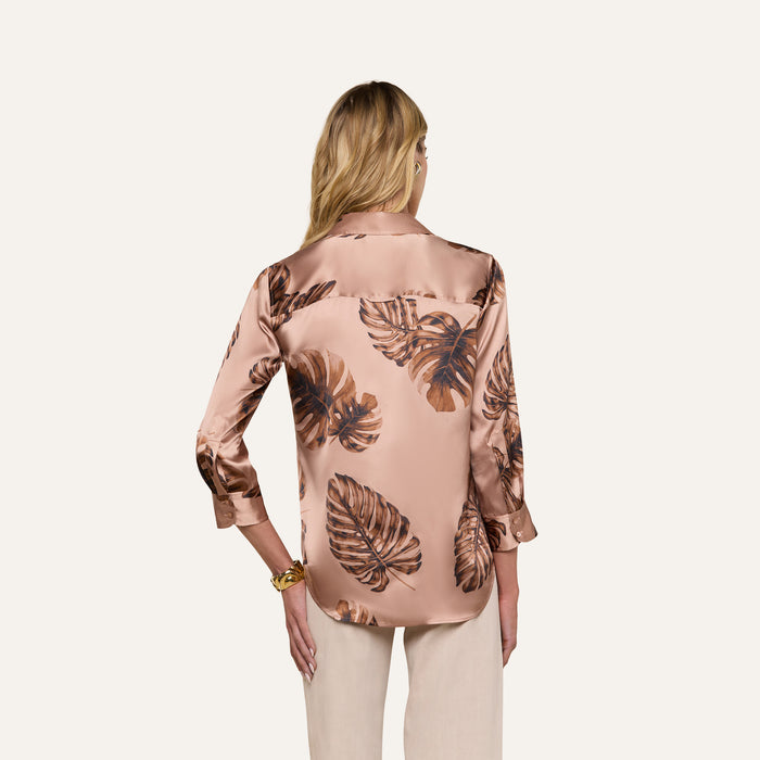 A woman with long blonde hair is seen from the back, wearing a shiny Dani Silk Blouse in light brown with a dark brown palm leaf print, paired with light beige pants, set against a plain light background.