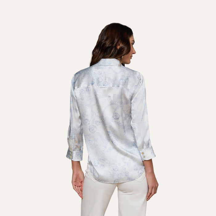 A woman with long brown hair, wearing the Dani Silk Blouse—a light blue patterned silk button-up shirt with rolled-up sleeves—and white pants, stands with her back to the camera against a plain background.