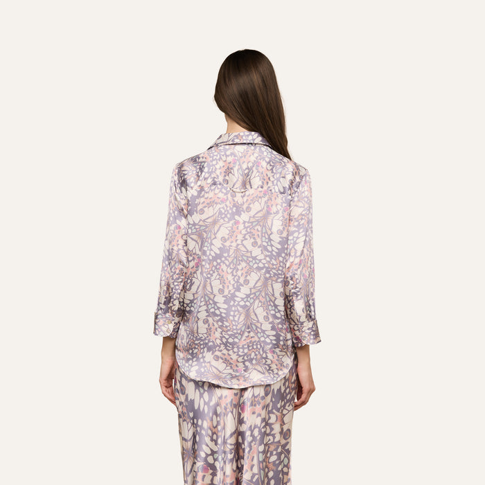A woman with long brown hair, seen from behind, wears the Dani Silk Blouse featuring a light purple and white floral pattern against an off-white background.