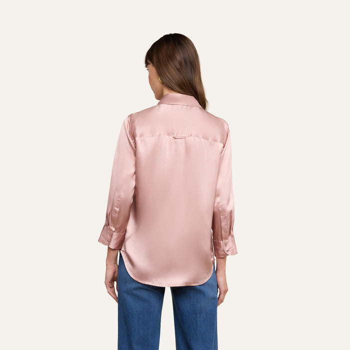 A woman stands with her back to the camera, wearing a Dani Silk Blouse in light pink, crafted from 100 silk charmeuse with long sleeves, paired with blue jeans, set against a plain white background.