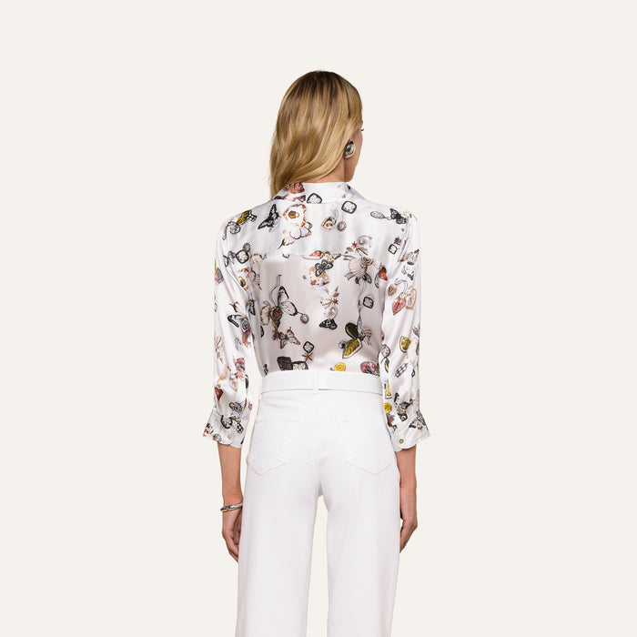 A woman with long blonde hair, dressed in the Dani Silk Blouse featuring a colorful print and paired with white pants, is shown from the back against a plain light background.