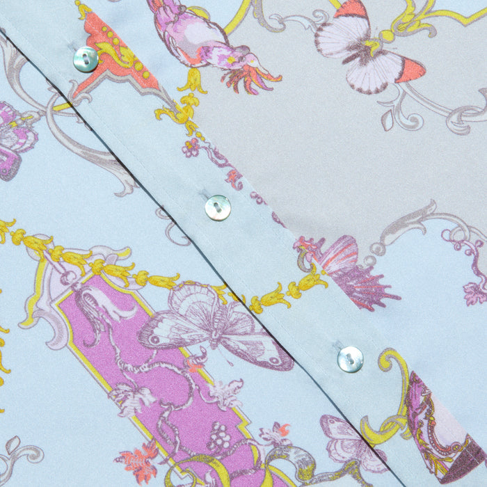 Close-up of the Dani Silk Blouse, a light blue patterned silk button-down shirt with shiny buttons, adorned with pink, purple, and yellow ornate designs featuring butterflies and decorative baroque toile print motifs.