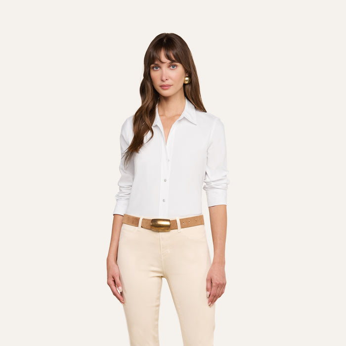 Wearing the Daniella Cotton-Blend Blouse, a woman with long brown hair stands against a plain, light background. The white three-quarter sleeve stretch cotton poplin blouse is tucked into beige pants with a tan belt. She looks slightly to the side with a relaxed expression.