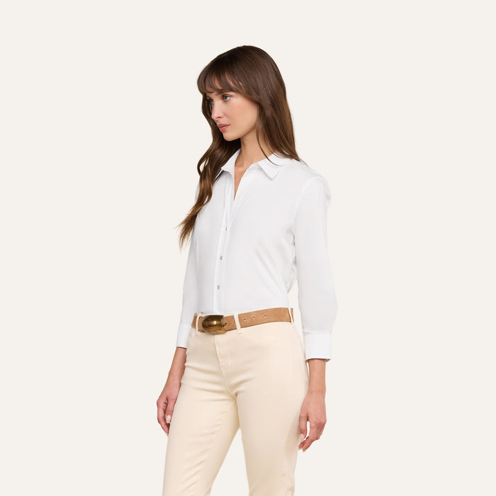 Wearing the Daniella Cotton-Blend Blouse in a relaxed fit, a woman stands against a plain light background, pairing the white stretch cotton poplin button-down shirt with beige pants and a tan belt. She looks to her left with her arms relaxed by her sides.