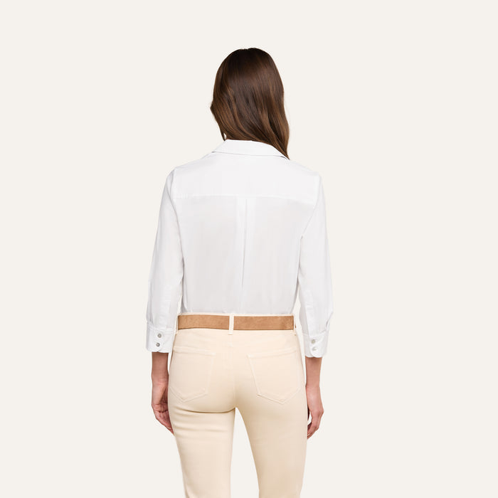 Woman standing with her back to the camera, wearing the Daniella Cotton-Blend Blouse—a relaxed fit white stretch cotton poplin long-sleeve shirt—tucked into high-waisted, light beige pants with a tan belt, against a plain off-white background.