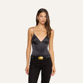A woman with long brown hair wears the Dara Stretch-Satin Bodysuit, featuring adjustable straps. She pairs it with black pants, a black belt with a large gold buckle, and gold earrings, standing against a plain, light background.