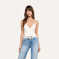 A woman with long brown hair wears the Dara Stretch Satin Bodysuit—a white sleeveless top with adjustable straps—paired with light blue jeans and a white belt. She is standing and looking at the camera against a plain light background.