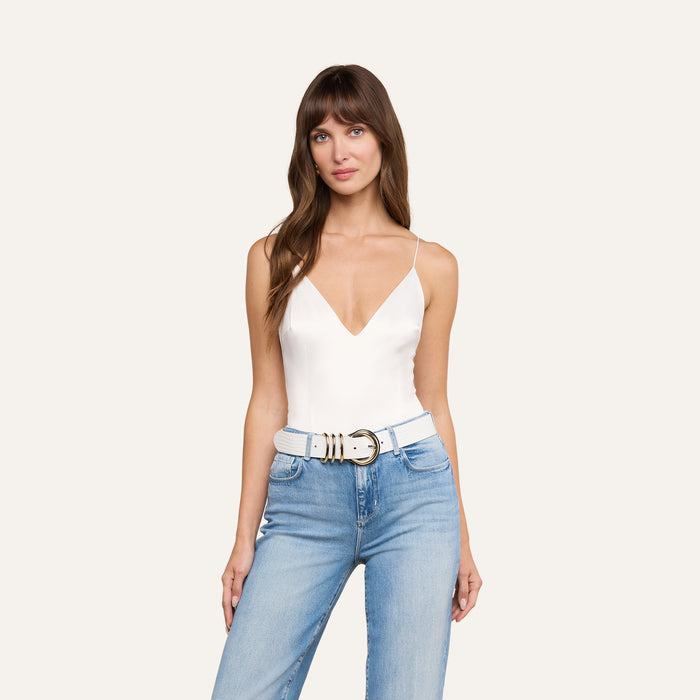 A woman with long brown hair wears the Dara Stretch Satin Bodysuit—a white sleeveless top with adjustable straps—paired with light blue jeans and a white belt. She is standing and looking at the camera against a plain light background.