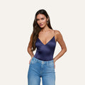 A woman with long brown hair wears the Dara Stretch-Satin Bodysuit in navy blue, featuring a plunge neckline and adjustable straps, paired with light blue jeans. She stands with her hands in her pockets against a plain, light background.