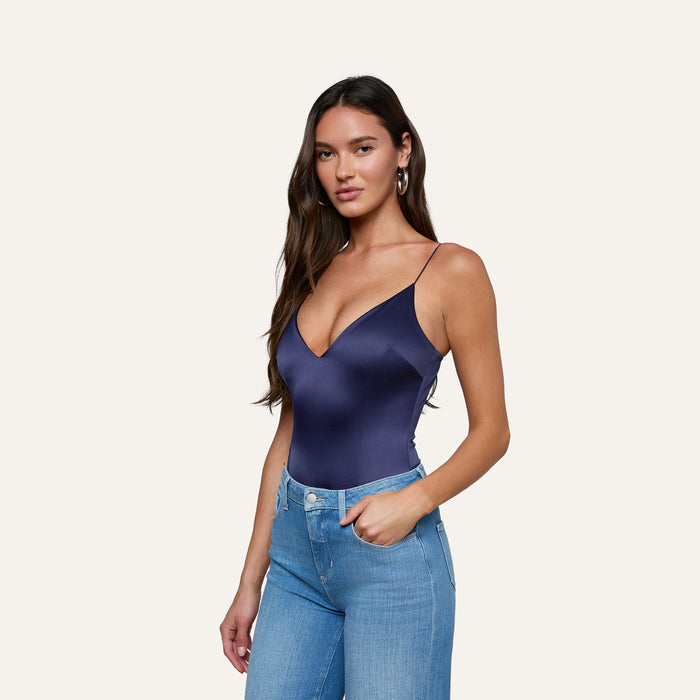 A woman with long brown hair wears the Dara Stretch-Satin Bodysuit in navy blue, featuring a plunge neckline, paired with light blue jeans and standing against a plain light background.