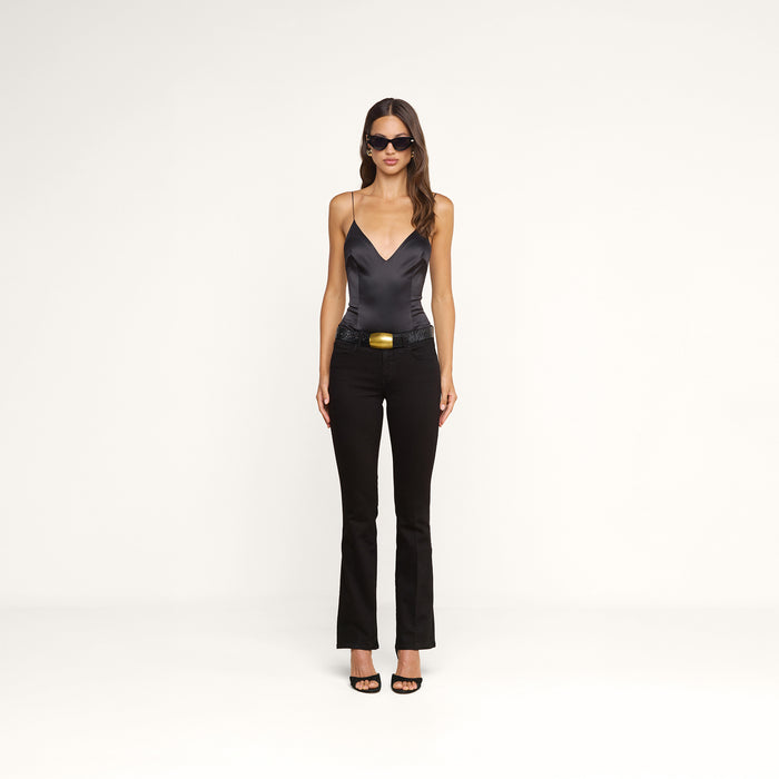 A woman stands against a plain white background, facing forward with her arms at her sides, wearing black sunglasses, the Dara Stretch-Satin Bodysuit with adjustable straps, black flared pants featuring a wide belt, and black heels.