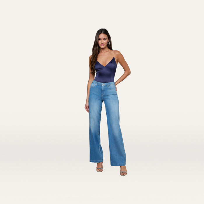 A woman wearing the Dara Stretch-Satin Bodysuit, styled with light blue high-waisted wide-leg jeans and open-toe heels, stands against a plain white background.