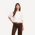A woman stands against a plain background wearing the Daria Oversized Cropped Tee in white, styled with brown pants and a brown belt featuring a gold buckle. She has long brown hair and rests her left hand on her hip.