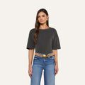 A woman with long brown hair is wearing a dark gray Daria Oversized Cropped Tee, blue jeans with a tan belt, and gold earrings, standing against a plain light background.