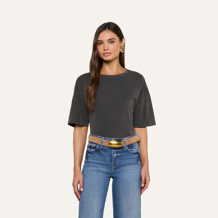 A woman with long brown hair is wearing a dark gray Daria Oversized Cropped Tee, blue jeans with a tan belt, and gold earrings, standing against a plain light background.