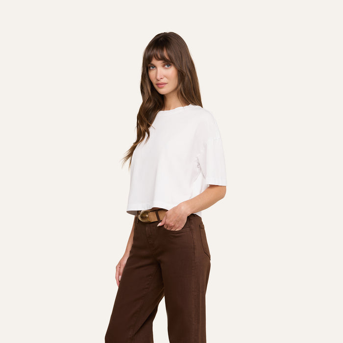 Woman with long brown hair wearing the Daria Oversized Cropped Tee in white and brown pants, standing with one hand in her pocket against a plain background.