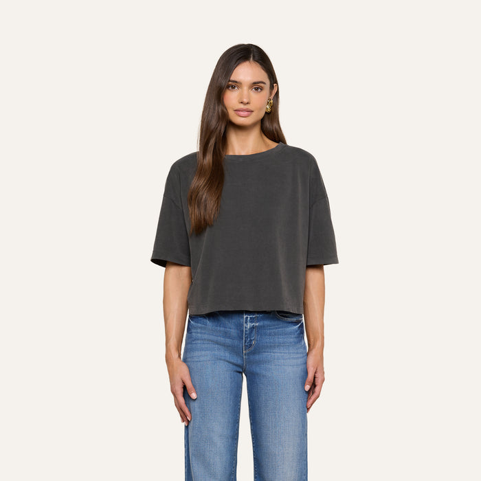 A woman faces forward against a plain off-white background, wearing the Daria Oversized Cropped Tee in black styled with light blue jeans. Her straight brown hair is worn simply, and gold hoop earrings complete her look.