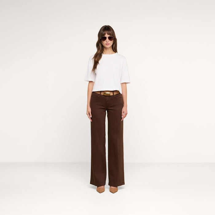 A woman wearing sunglasses, the Daria Oversized Cropped Tee in white made from Supima cotton, brown wide-leg pants with a belt, and tan heels stands against a plain white background.