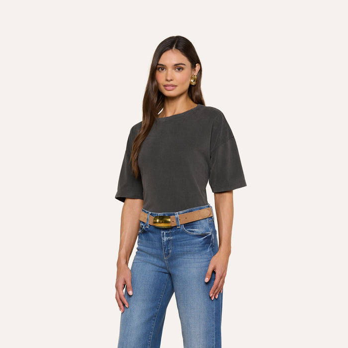 A woman with long brown hair and gold earrings stands against a plain light background, wearing the Daria Oversized Cropped Tee in dark gray. The short-sleeve t-shirt is styled in an oversized fit, tucked into blue jeans with a wide belt.