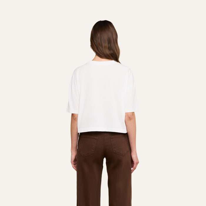 A person with long brown hair, seen from behind against a plain light background, wears the Daria Oversized Cropped Tee in white paired with brown pants.