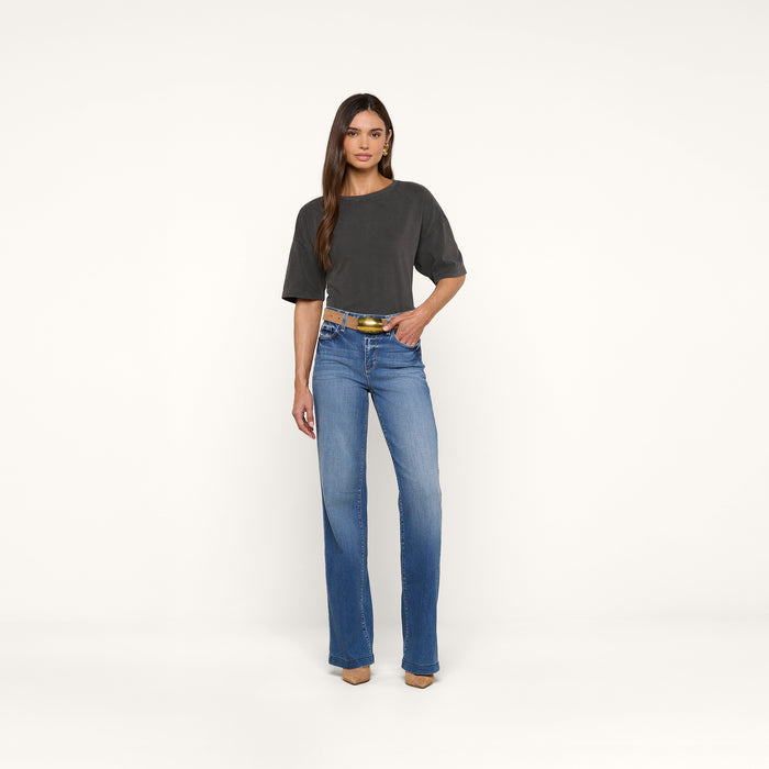 A woman stands against a plain white background wearing the Daria Oversized Cropped Tee in dark gray with blue jeans, a yellow belt, and tan shoes. She has long, straight hair and one hand in her pocket.