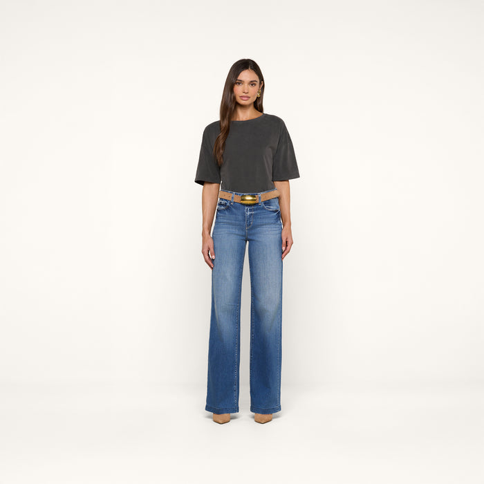 A woman stands facing forward, wearing the Daria Oversized Cropped Tee in dark gray with an oversized fit, blue wide-leg jeans, a black belt with a gold buckle, and beige pointed shoes against a plain white background.