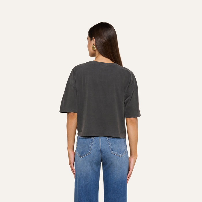 A woman stands facing away, wearing the Daria Oversized Cropped Tee in dark gray, made from Supima cotton, paired with blue jeans. Her straight brown hair falls over her shoulders, and she accessorizes with gold hoop earrings. The background is plain and light-colored.