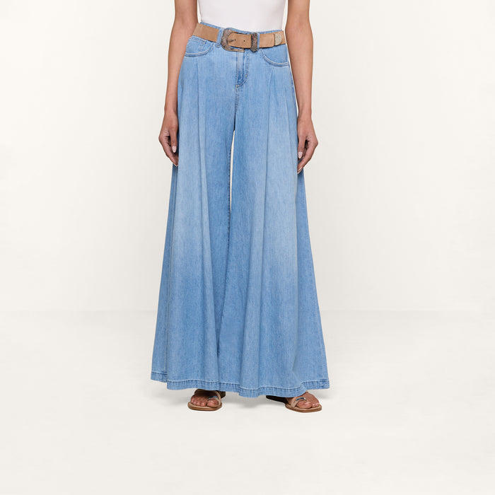 Wearing the Dariela Wide-Leg Jean in light blue, paired with a brown belt, sandals, and a white sleeveless top, this person stands against a plain white background.