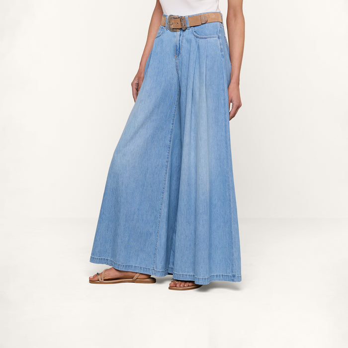 A person wearing the Dariela Wide-Leg Jean in light blue denim with pleats, paired with a brown belt, white top, and tan sandals stands against a plain white background.