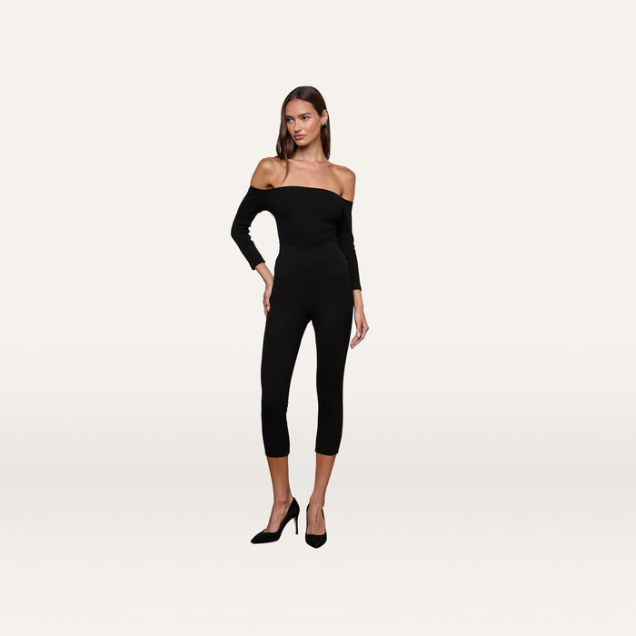 A woman stands against a plain light background, looking slightly to her left with one hand on her hip to accentuate the flattering fit of the Dee Capri Pant, styled here as a black off-the-shoulder fitted jumpsuit in stretch ponte with capri-length legs and paired with black heels.