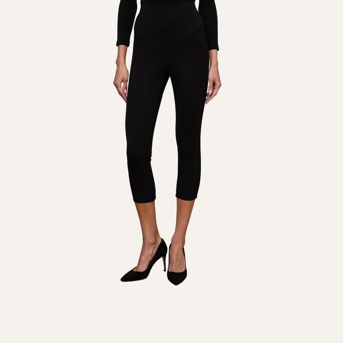 A person wearing the Dee Capri Pant in black along with black high-heeled shoes stands against a plain, light background. Only the legs and lower torso are visible, showcasing the pant’s flattering fit.