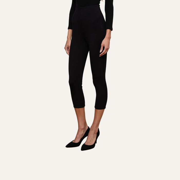 A woman is shown from the shoulders down, standing against a plain light background, wearing the Dee Capri Pant in black stretch ponte with black heels. The flattering fit of these capri pants is prominently displayed.