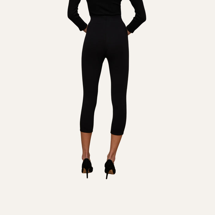 A person wearing the Dee Capri Pant in black, styled with a black long-sleeve top and black heels, stands facing away on a plain light background.