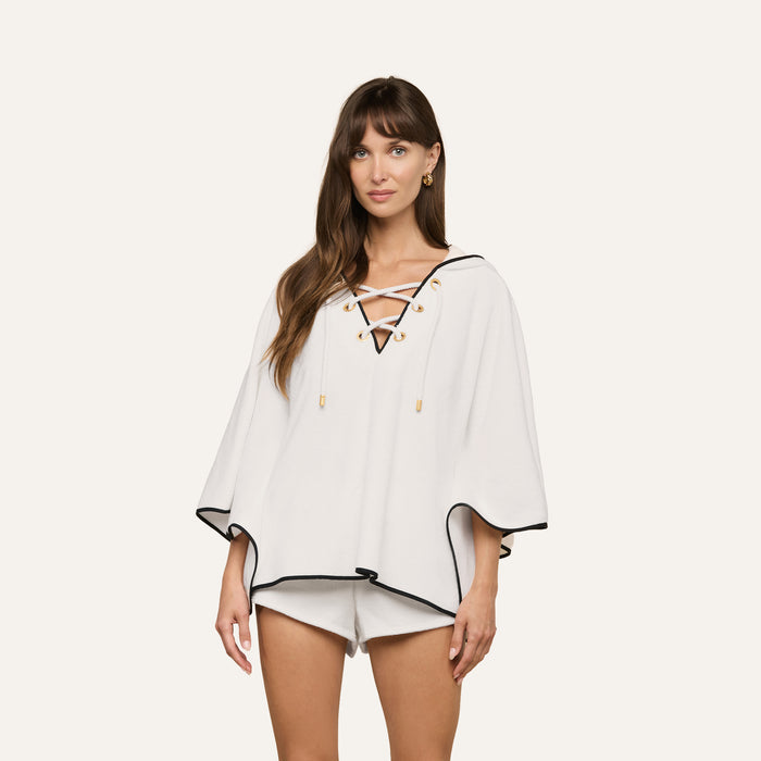 A woman stands against a plain light background, radiating spa-like energy in the Demeter Cover-Up Poncho—a white, loose-fitting poncho featuring black trim and a lace-up front detail. She styles it with crisp white shorts for an effortlessly relaxed look.