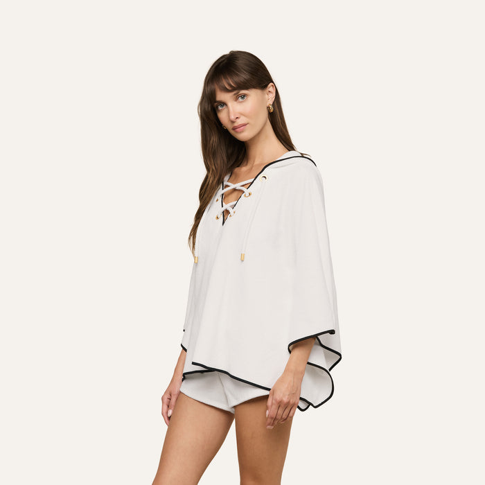 A woman models the Demeter Cover-Up Poncho—a white terrycloth poncho top featuring a lace-up front and black trim—paired with white shorts, set against a plain light background. This outfit exudes effortless spa-inspired relaxation.