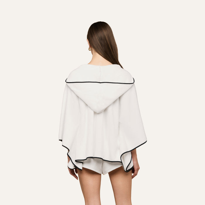 A person with long brown hair is shown from the back wearing the Demeter Cover-Up Poncho—a white terrycloth cover-up with black trim—paired with matching white shorts, set against a plain light background.