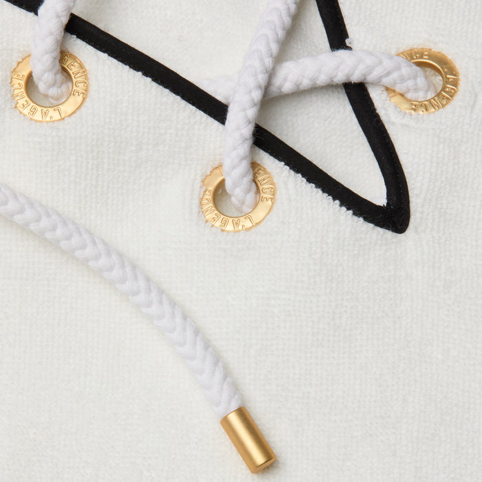 Closeup of white braided drawstrings threaded through gold metal eyelets on the Demeter Cover-Up Poncho—a white textured terrycloth piece featuring a single black stripe and a gold-capped drawstring end.