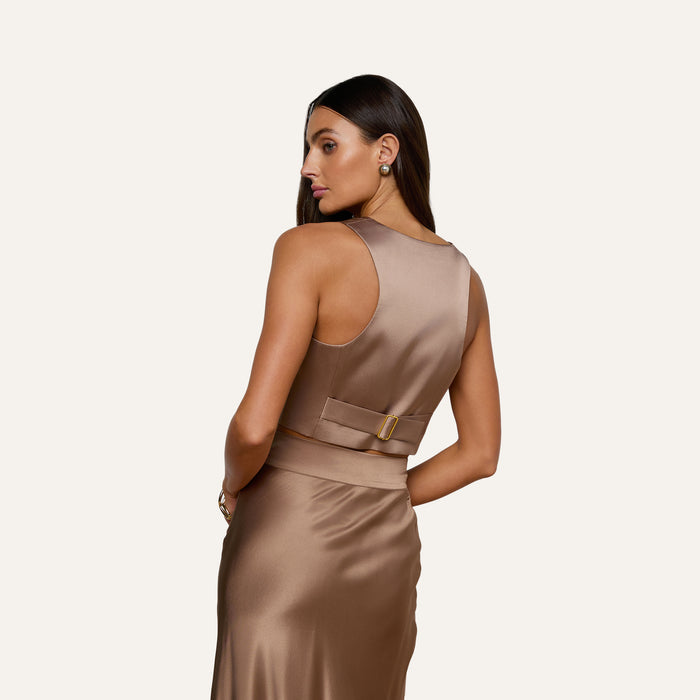 A woman with long brown hair stands against a plain light background, looking over her shoulder while wearing the Devony Silk Vest—a sleeveless, light brown satin top featuring a tailored evening silhouette and a belt detail at the back.