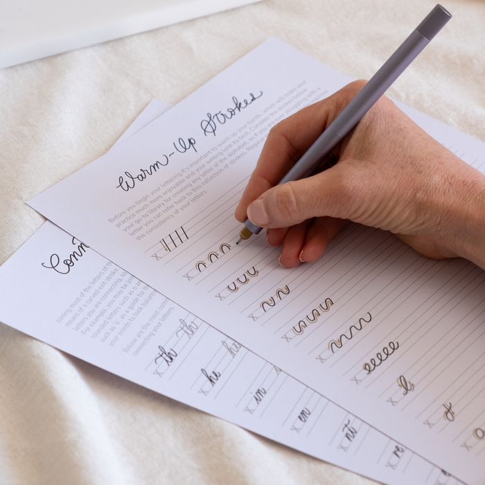A hand using a Lilac coloured Lumos pen with fineliner tip to trace warm-up strokes from the Monoline Lettering Guide by Tom’s Studio. The printed sheet includes practice lines and curved stroke patterns.
