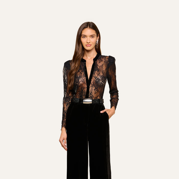 A woman with long brown hair wearing the black Dorothea Velvet Lace Blouse and high-waisted black pants, one hand in her pocket, stands against a plain light background.