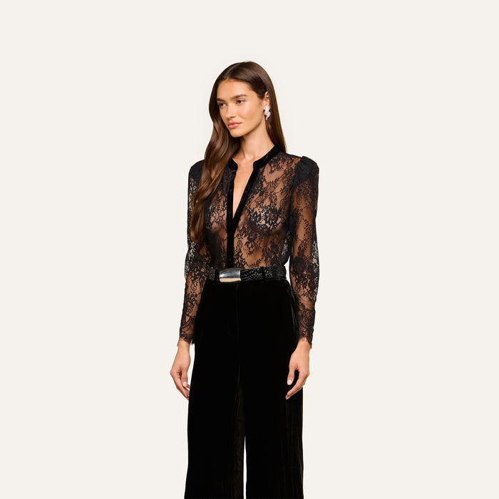A woman with long brown hair wears the Dorothea Velvet Lace Blouse, paired with black high-waisted pants, a black belt with a silver buckle, and drop earrings, standing against a plain light background.