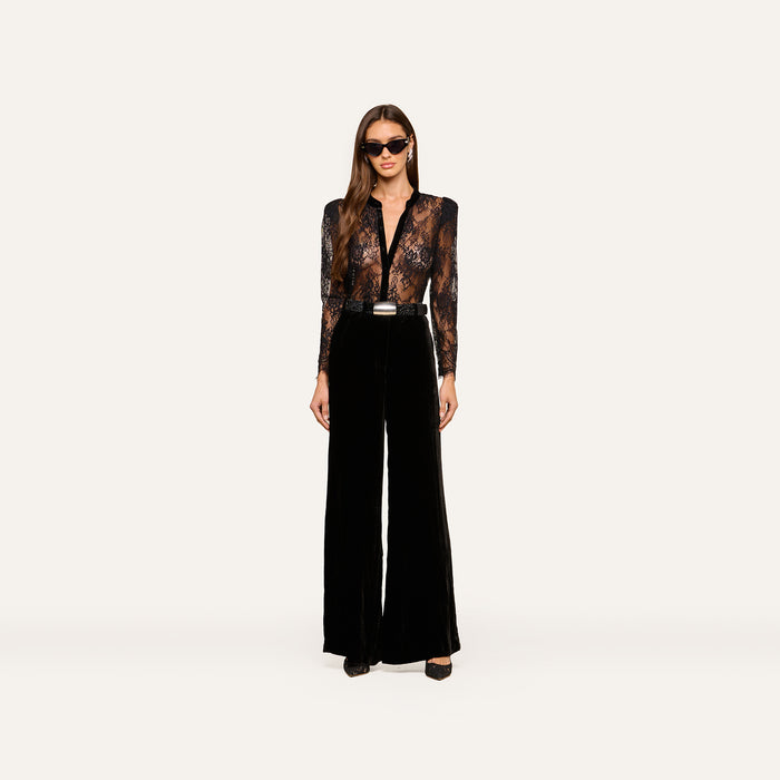 A woman wearing sunglasses, the Dorothea Velvet Lace Blouse—featuring sheer Chantilly lace with velvet seams—and high-waisted black wide-leg pants stands facing forward against a plain white background.