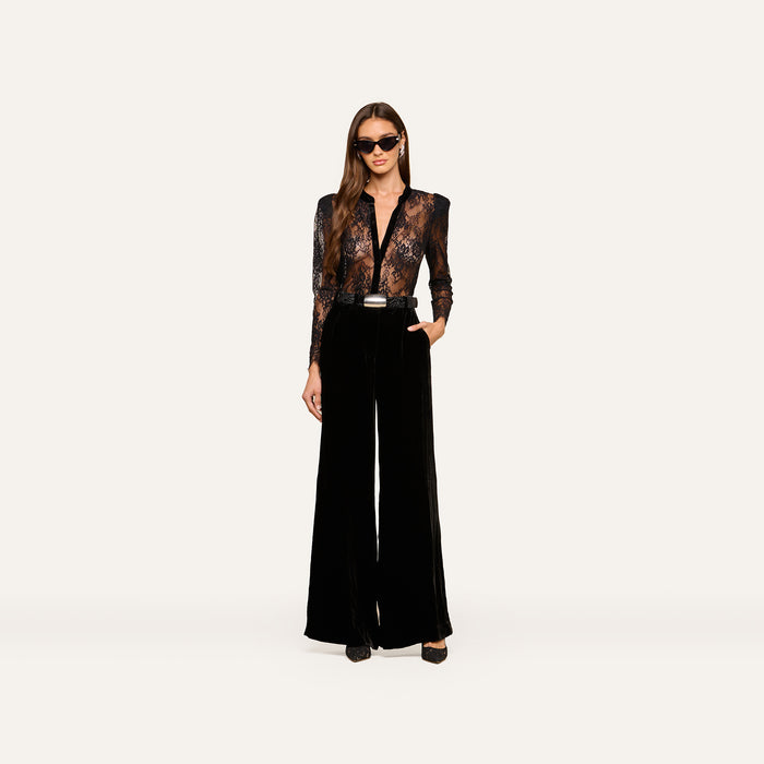 Wearing the Dorothea Velvet Lace Blouse, a woman in sunglasses stands confidently with one hand in her pocket, showcasing its sheer Chantilly lace long sleeves tucked into high-waisted, wide-leg black velvet pants with elegant velvet seams and finished with black shoes.