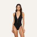 A woman models the Dorrie Plunge One-Piece Swimsuit, featuring a deep V-neckline and a gold front zipper, standing against a plain light background.